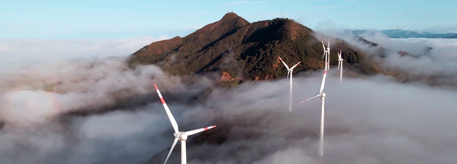 Luftaufnahme von einer Drohne eines Windparks über den Wolken mit einem Berg im Hintergrund und blauem Himmel