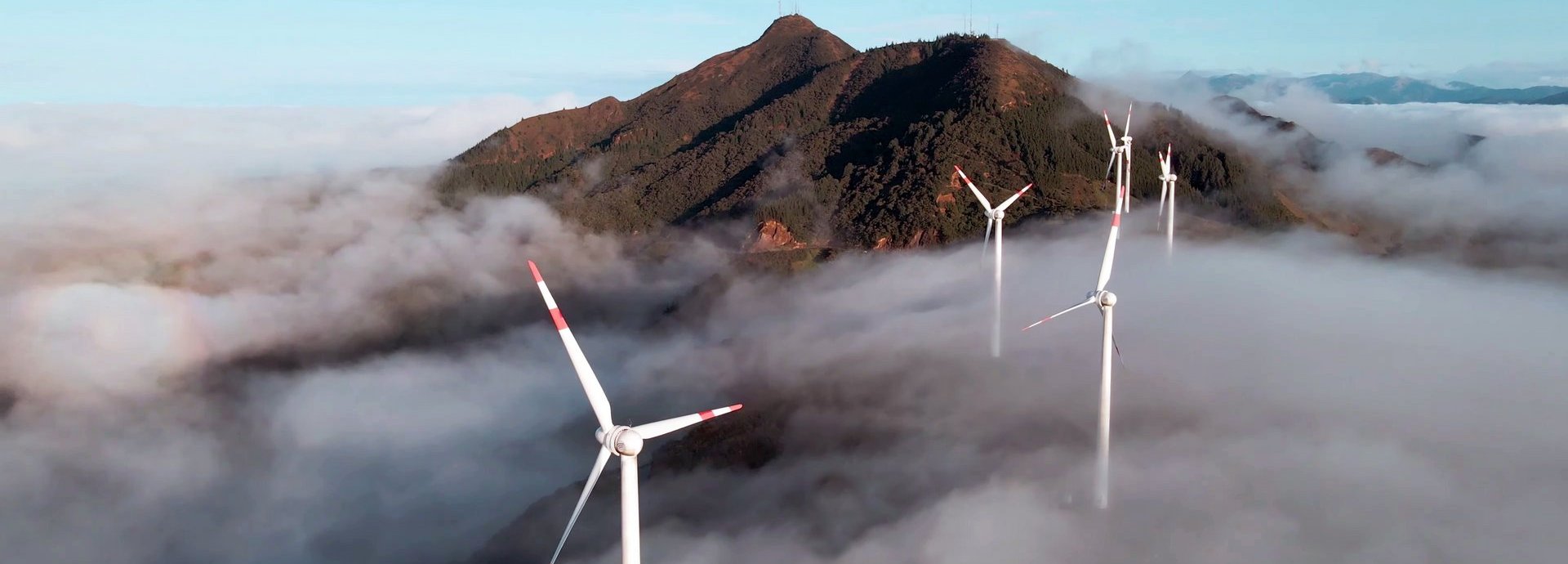 Luftaufnahme von einer Drohne eines Windparks über den Wolken mit einem Berg im Hintergrund und blauem Himmel