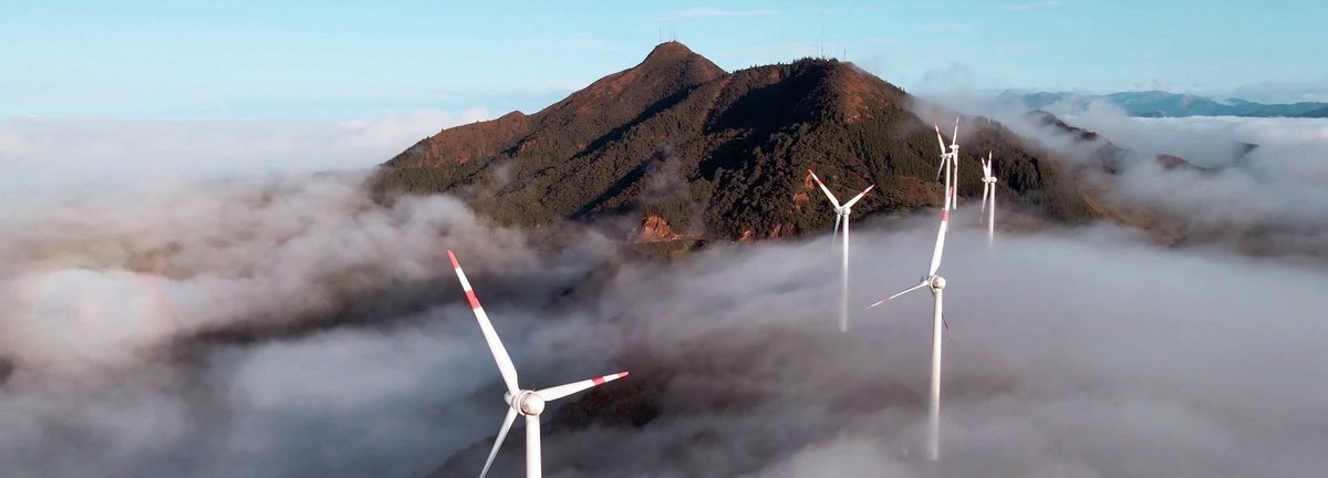 Luftaufnahme von einer Drohne eines Windparks über den Wolken mit einem Berg im Hintergrund und blauem Himmel