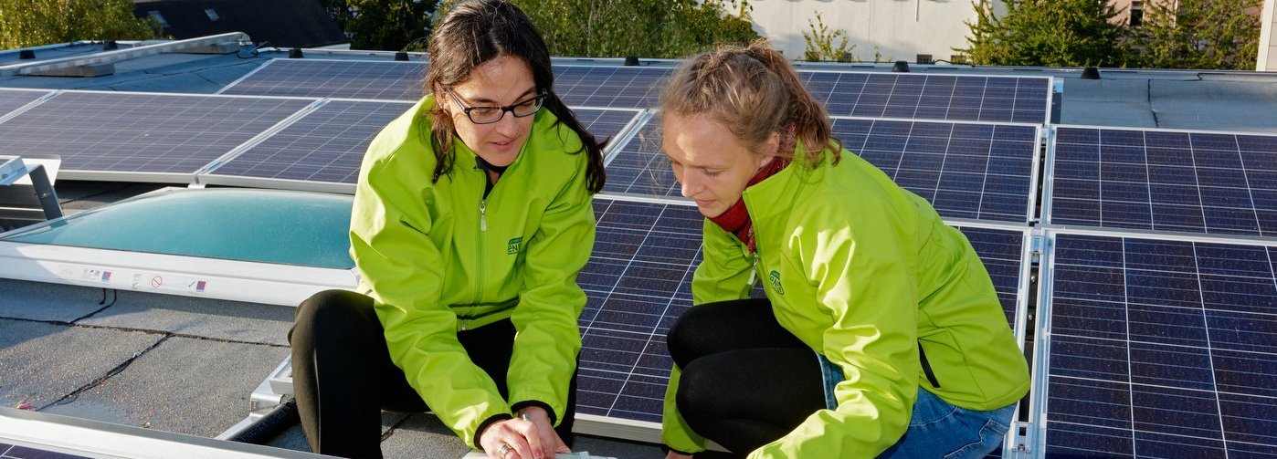 Zwei Forscherinnen in gelben Jacken hocken an Solarkollektoren und schreiben Notizen auf