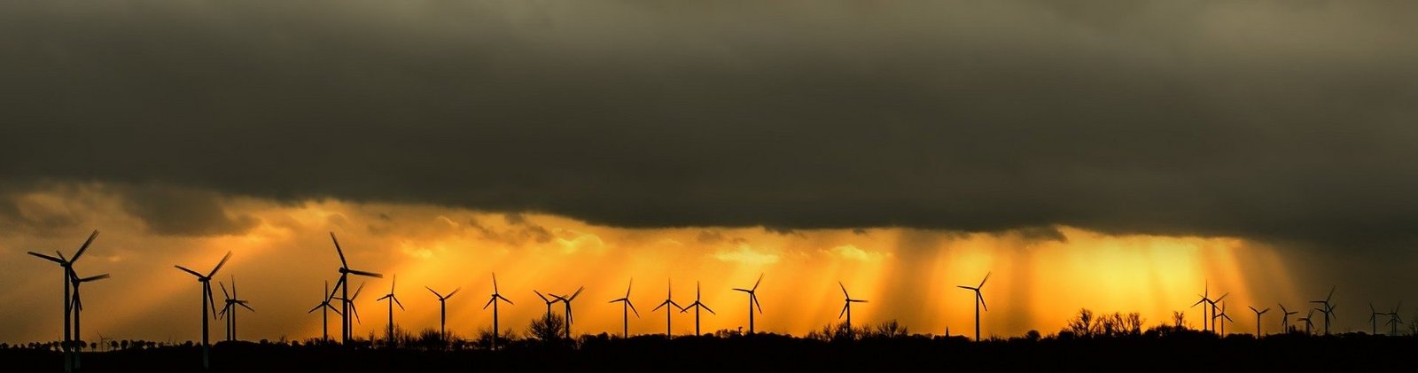 dunkler bewölkter Himmel aus dem die Sonne ausbricht und in orange eine Reihe von Windrädern am Horizont anleuchtet