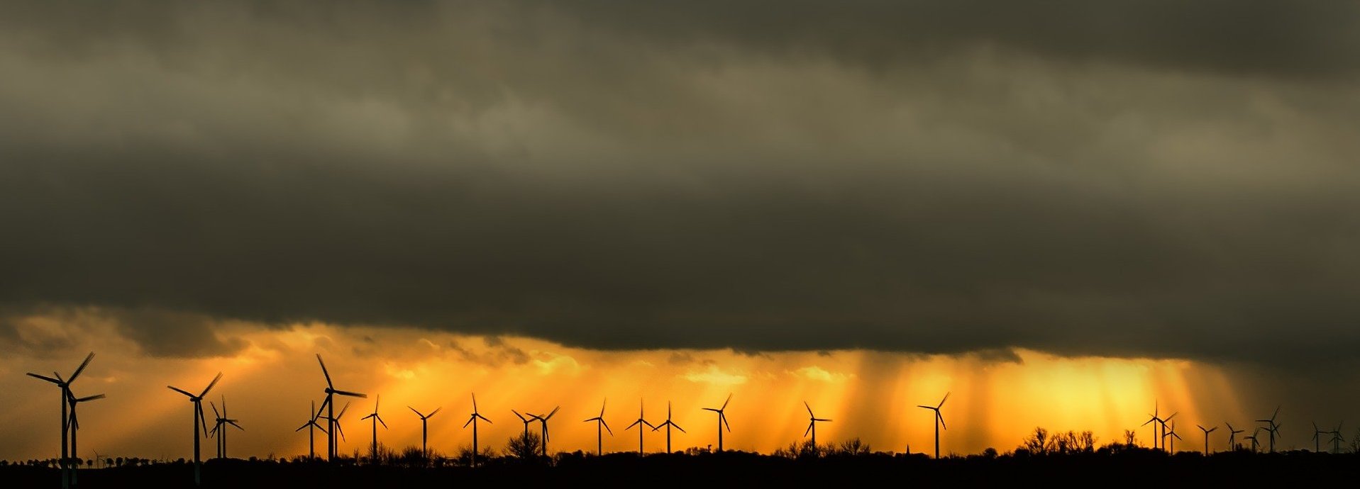 dunkler bewölkter Himmel aus dem die Sonne ausbricht und in orange eine Reihe von Windrädern am Horizont anleuchtet