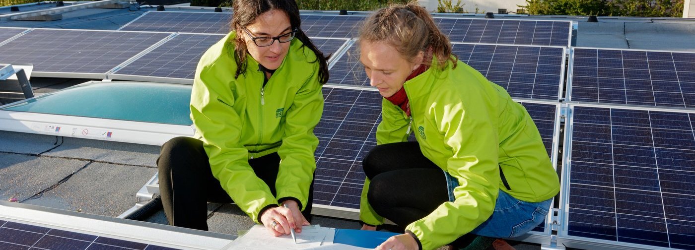 Zwei Mitarbeiterinnen der Green Planet Energy eG mit grünen Jacken auf einem Dach bei der Untersuchung von Solarzellen