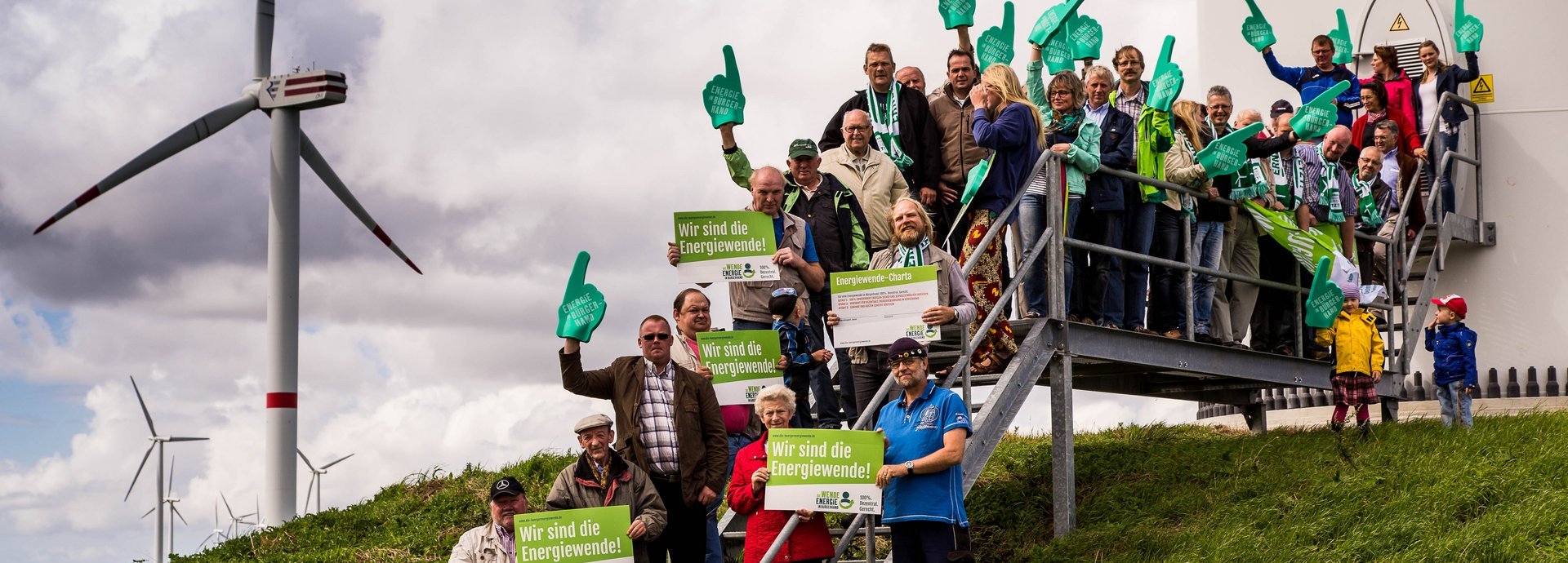 Gruppe von Menschen steht auf einer Wiese an der Metalltreppe vor einem Windrad mit Wir sind die Energiewende Schildern 