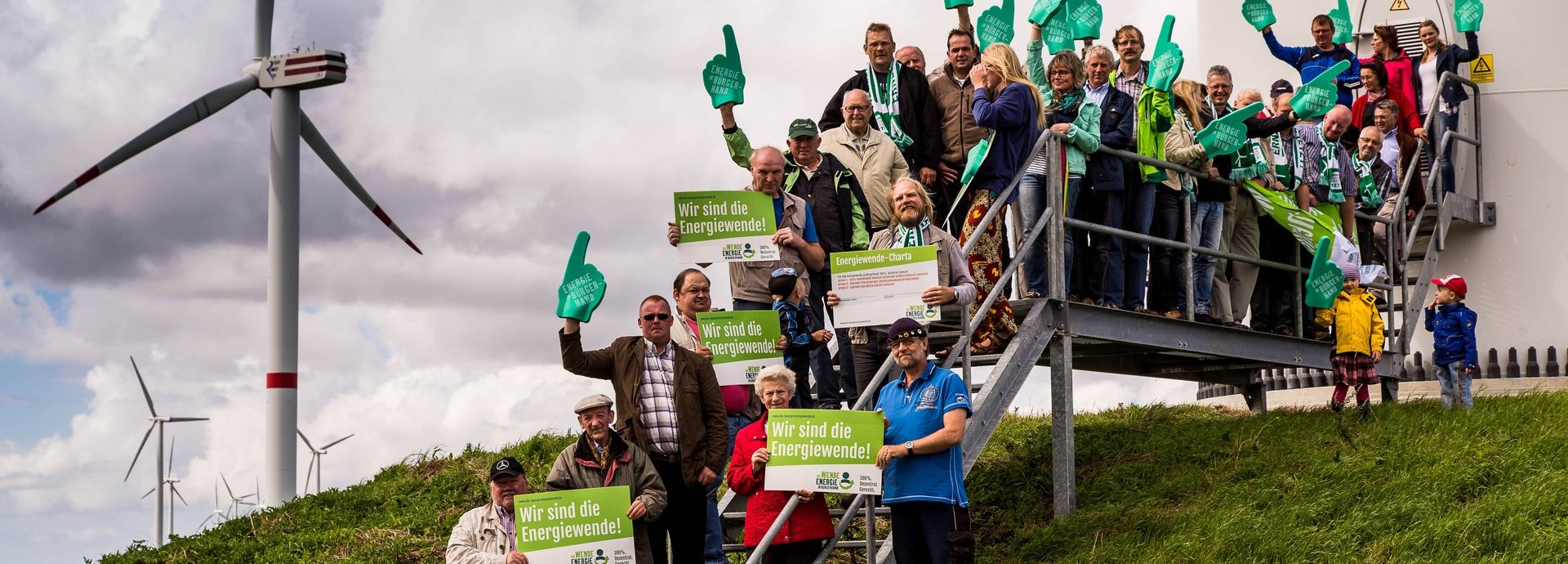 Gruppe von Menschen steht auf einer Wiese an der Metalltreppe vor einem Windrad mit Wir sind die Energiewende Schildern 