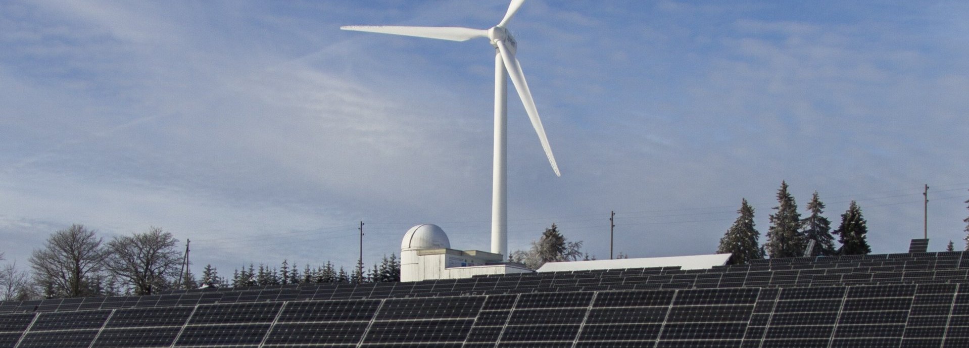 Solarkollektoren auf einer Wiese mit Schnee, Bäume und ein Windrad im Hintergrund