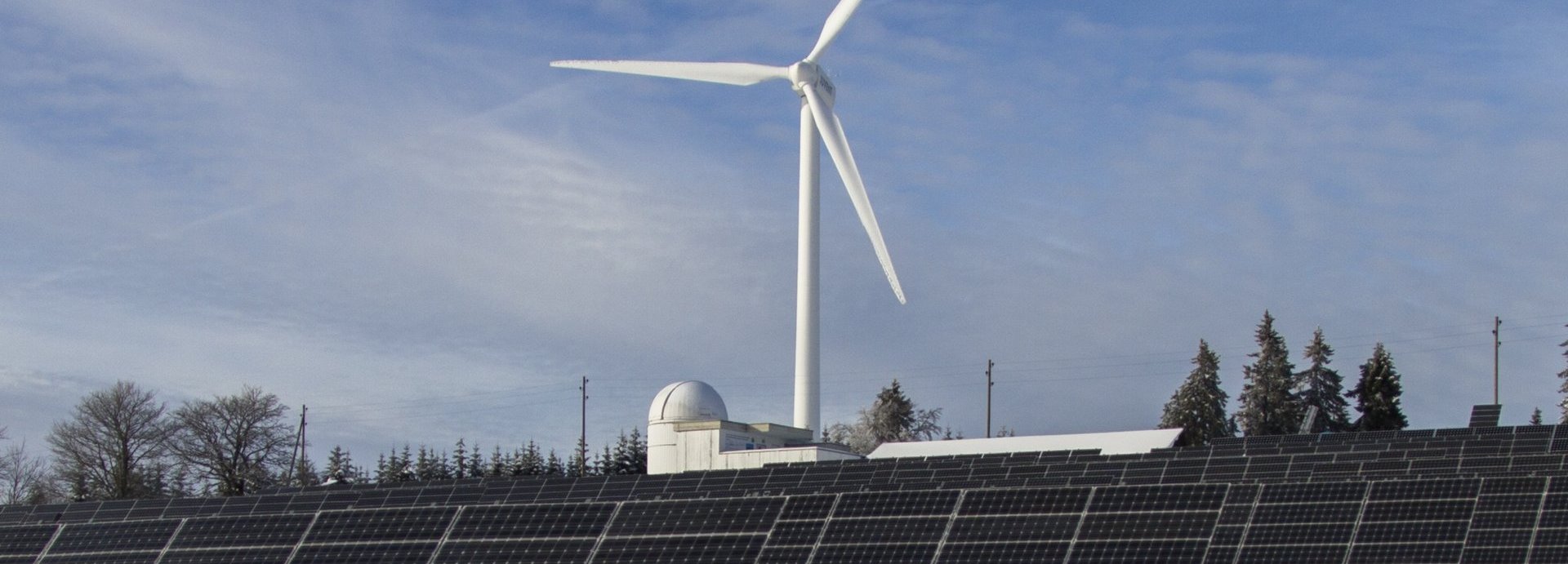 Solarkollektoren auf einer Wiese mit Schnee, Bäume und ein Windrad im Hintergrund