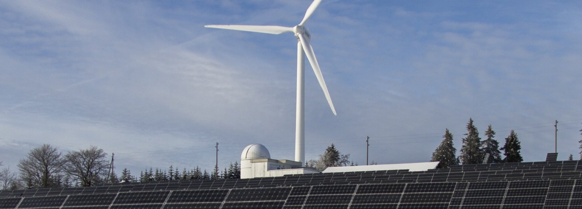 Solarkollektoren auf einer Wiese mit Schnee, Bäume und ein Windrad im Hintergrund