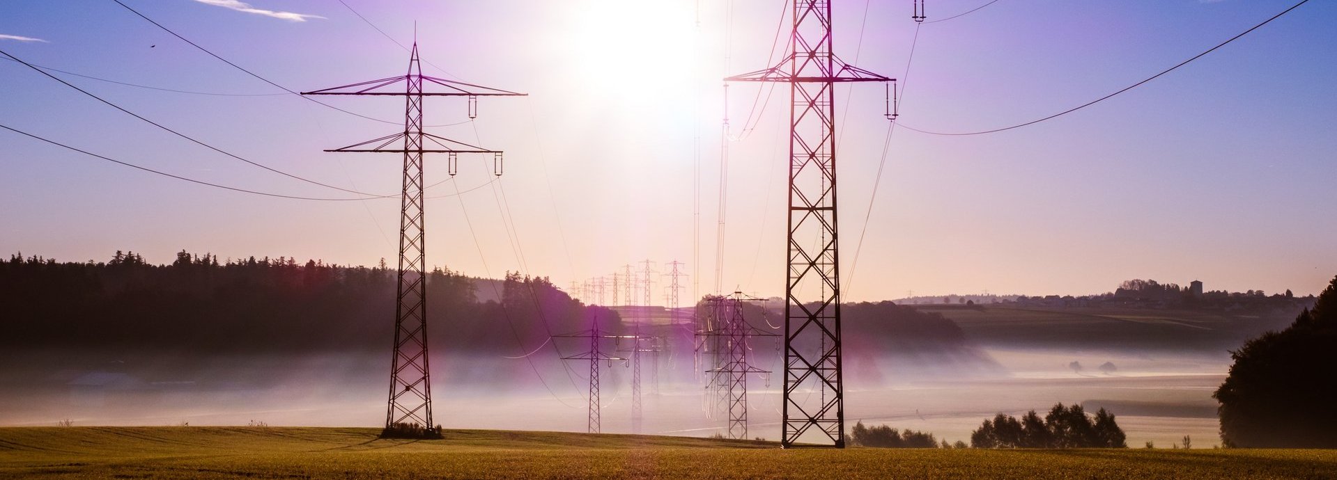 Stromtrasse vor dunkelblauem Hintergrund und Sonne auf einem nebligen Feld