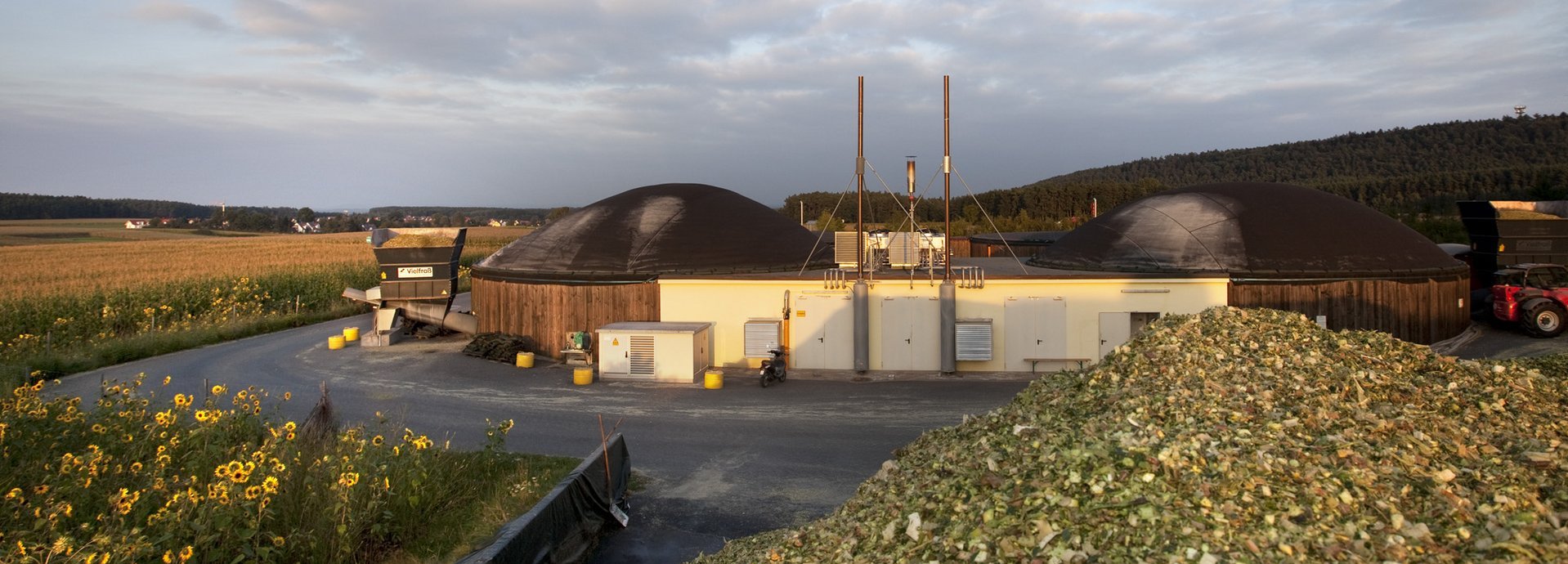 Beschreibung:Biogasanlage an einer Straße. Davor ein Berg aus Biomaterial zur Verbrennung. Auf der anderen Straßenseite ein Sonnenblumenfeld.
