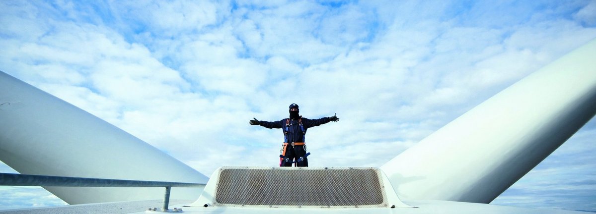 Monteur steht mit ausgebreiteten Armen auf der Gondel einer Windenergieanlage