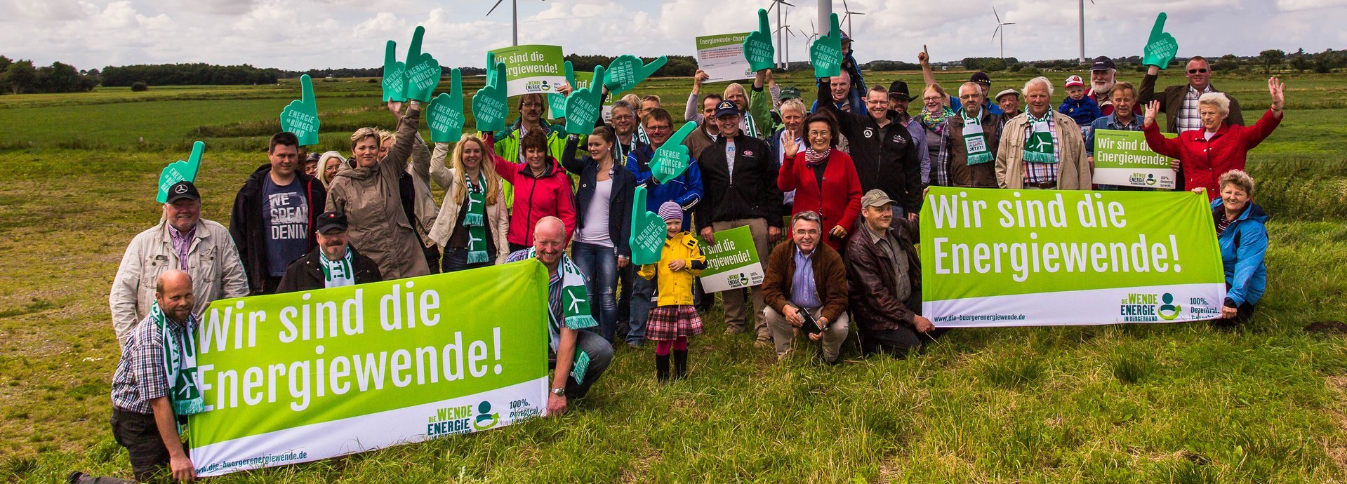 Menschengruppe mit Wir sind Energiewende Schildern auf einem Feld vor Windrädern