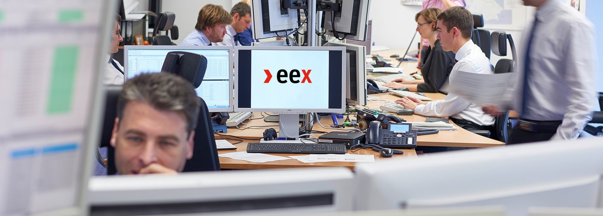 Mehrere Menschen in Business-Kleidung arbeiten in einem Büro an Rechnern. Auf einem Bildschirm in der Mette ist das >eex Logo