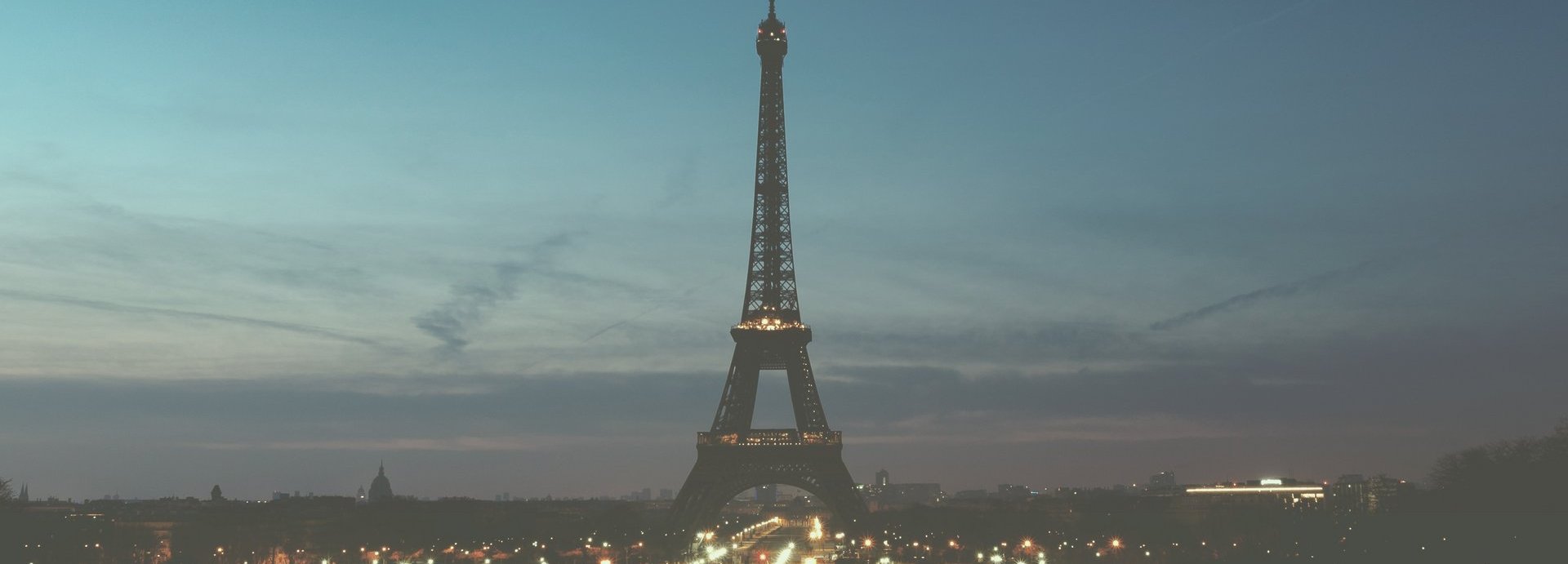 Fernaufnahme des Eiffelturms vor einem halb hellblauem halb dunkelblauem Himmel. Am Fuß des Eiffelturms beleuchtete Straßen