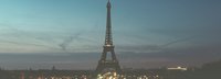 Fernaufnahme des Eiffelturms vor einem halb hellblauem halb dunkelblauem Himmel. Am Fuß des Eiffelturms beleuchtete Straßen