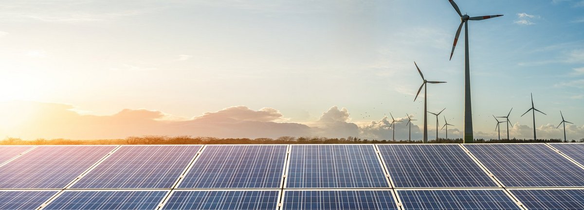 Windräder vor einem hellblauen Himmel mit tiefen Wolken und Solarkollektoren im Vordergrund