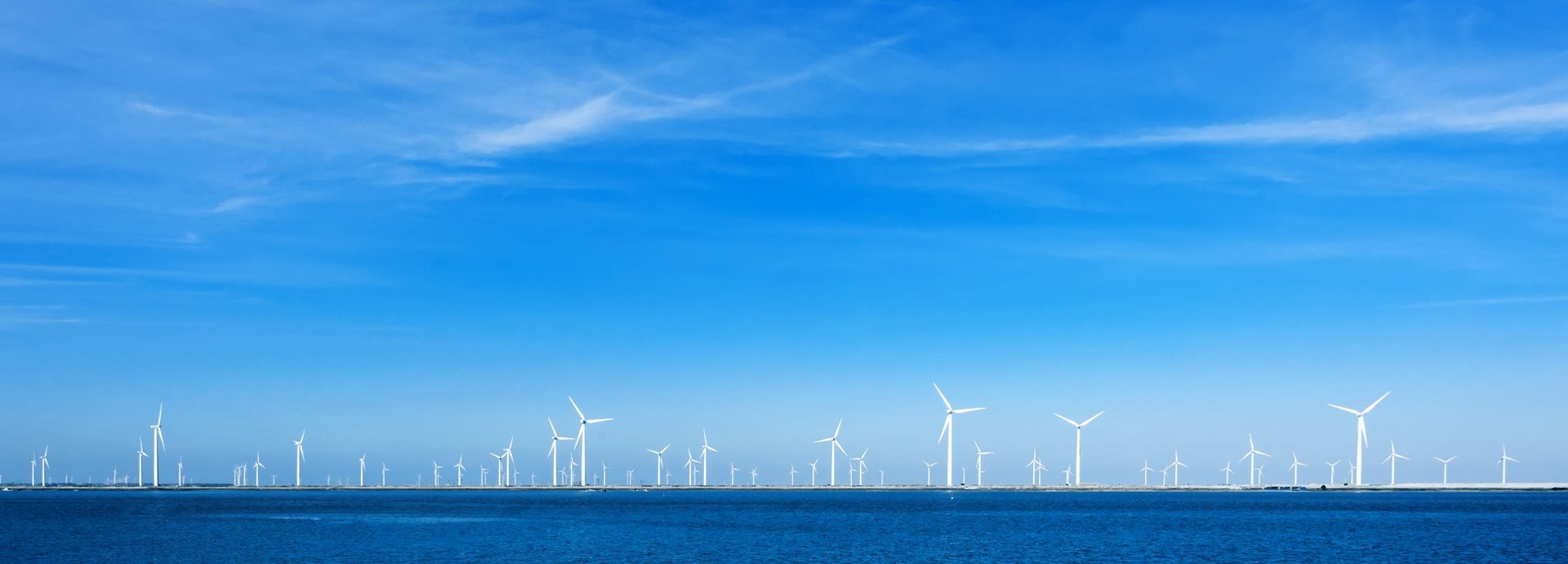 Viele Offshore Windräder am Horizont umgeben von blauem wolkenfreiem Himmel und dem Meer