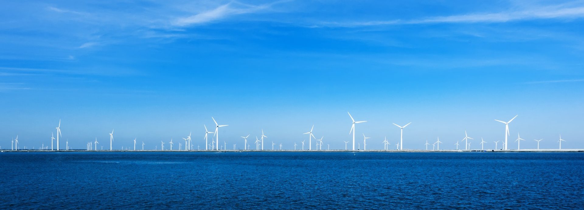 Viele Offshore Windräder am Horizont umgeben von blauem wolkenfreiem Himmel und dem Meer