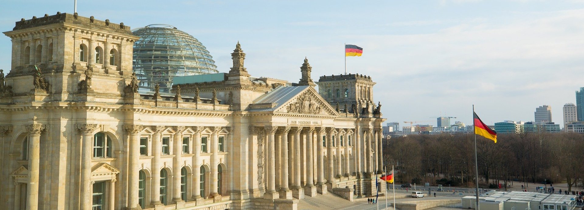 Weite Ansicht auf den Bundestag mit zwei wehenden deutschen Flaggen vor einem leicht bewölkten Himmel
