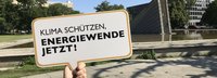 Arm hält Schild mit "Klima schützen, Energiewende jetzt" hoch. Im Hintergrund ein Baum und ein von Wasser umgebenes Gebäude