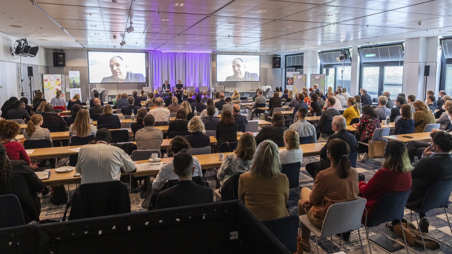 Global Business Scout Summit volle Veranstaltung Menschen Sitzen an Tischen und schauen auf zwei Leinwände an denen ein Mann spricht 