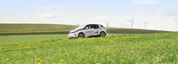 Elektroauto fährt auf einer grünen Wiese mit hügeliger Landschaft und Windrädern im Hintergrund