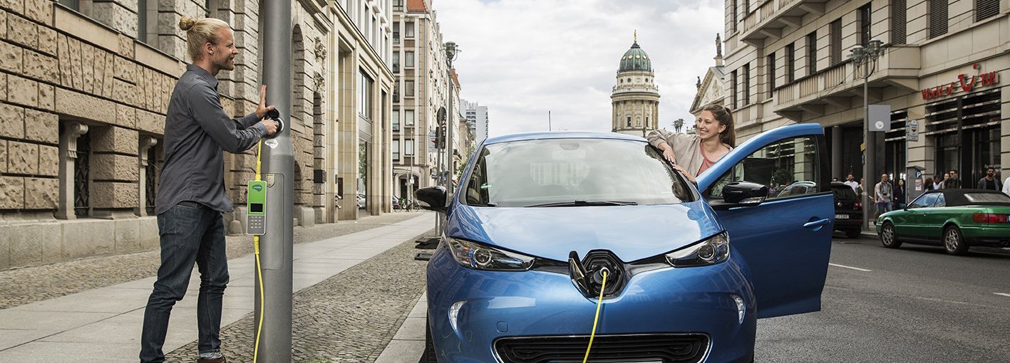 Mann lädt in der Stadt sein blaues Elektroauto an einer Laterne während lächelnde Frau an der Autotür wartet