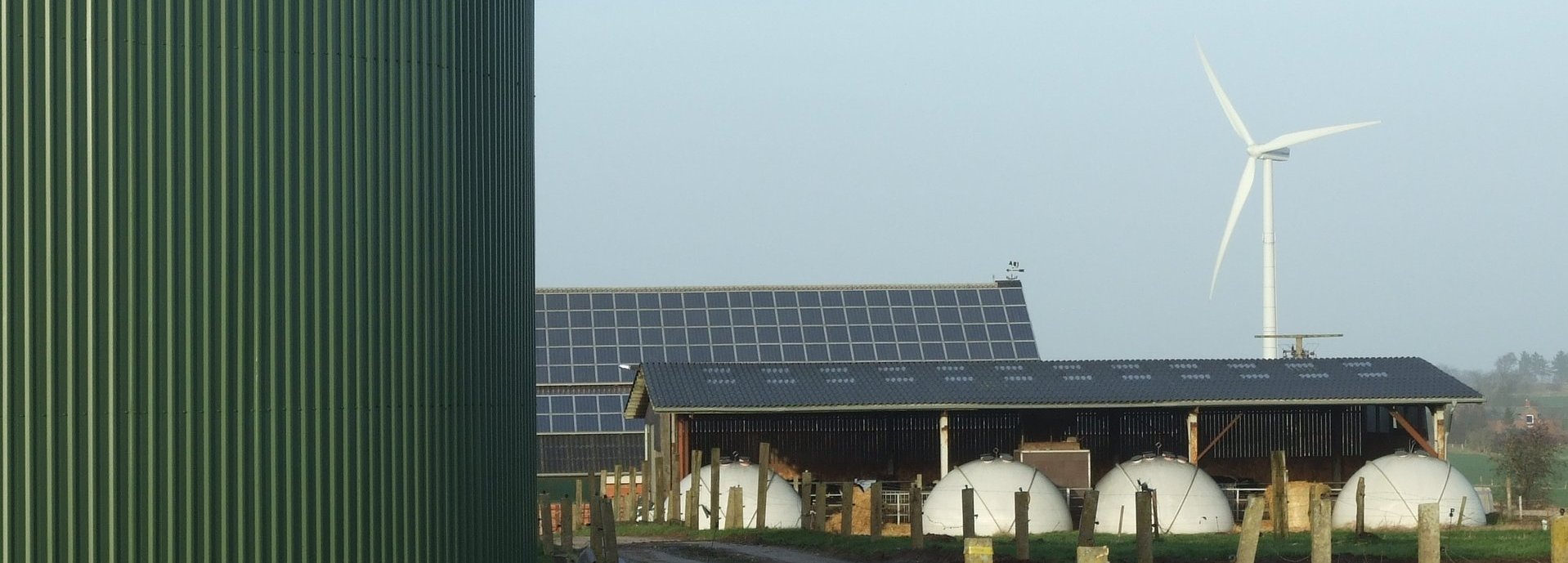 Nahaufnahme einer Biogasanlage. Im Hintergrund eine Tieranlage, Solarkollektoren und ein Windrad am Horizont