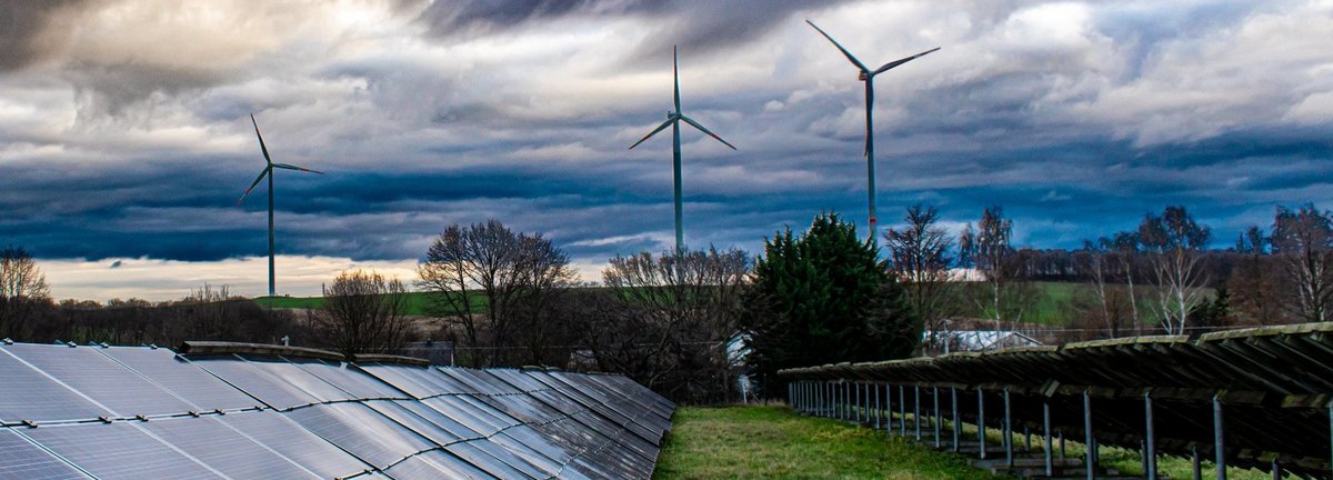 Reihen von Solarkollektoren auf einem Feld. Im Hintergrund Windräder. Der Himmel ist wolkenbehangen und dunkelblau.