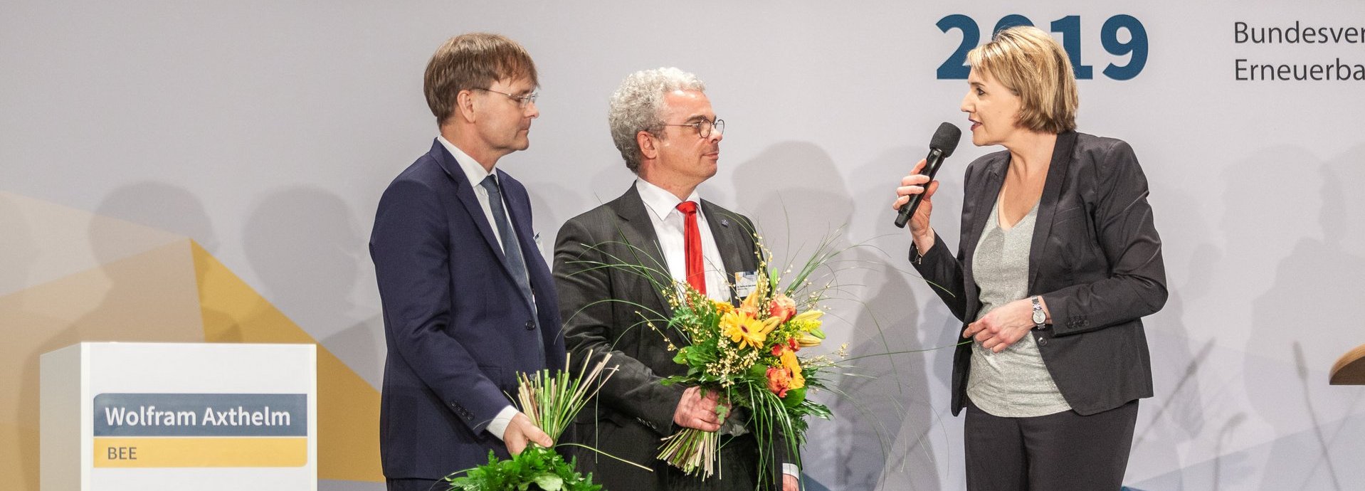 BEE Präsidentin Simone Peter spricht zu BEE Geschäftsführern Wolfram Axthelm und Claudius da Costa Gomez, die Blumensträuße in den Händen halten