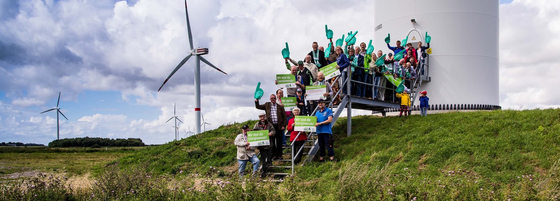 Menschengruppe mit Wir sind die Energiewende Schildern auf den Treppen vor einem Windrad