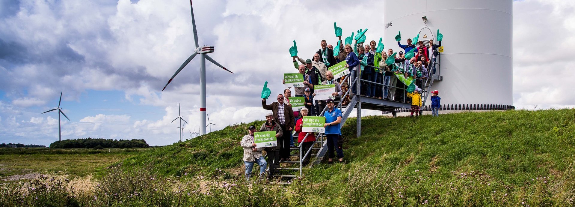 Menschengruppe mit Wir sind die Energiewende Schildern auf den Treppen vor einem Windrad
